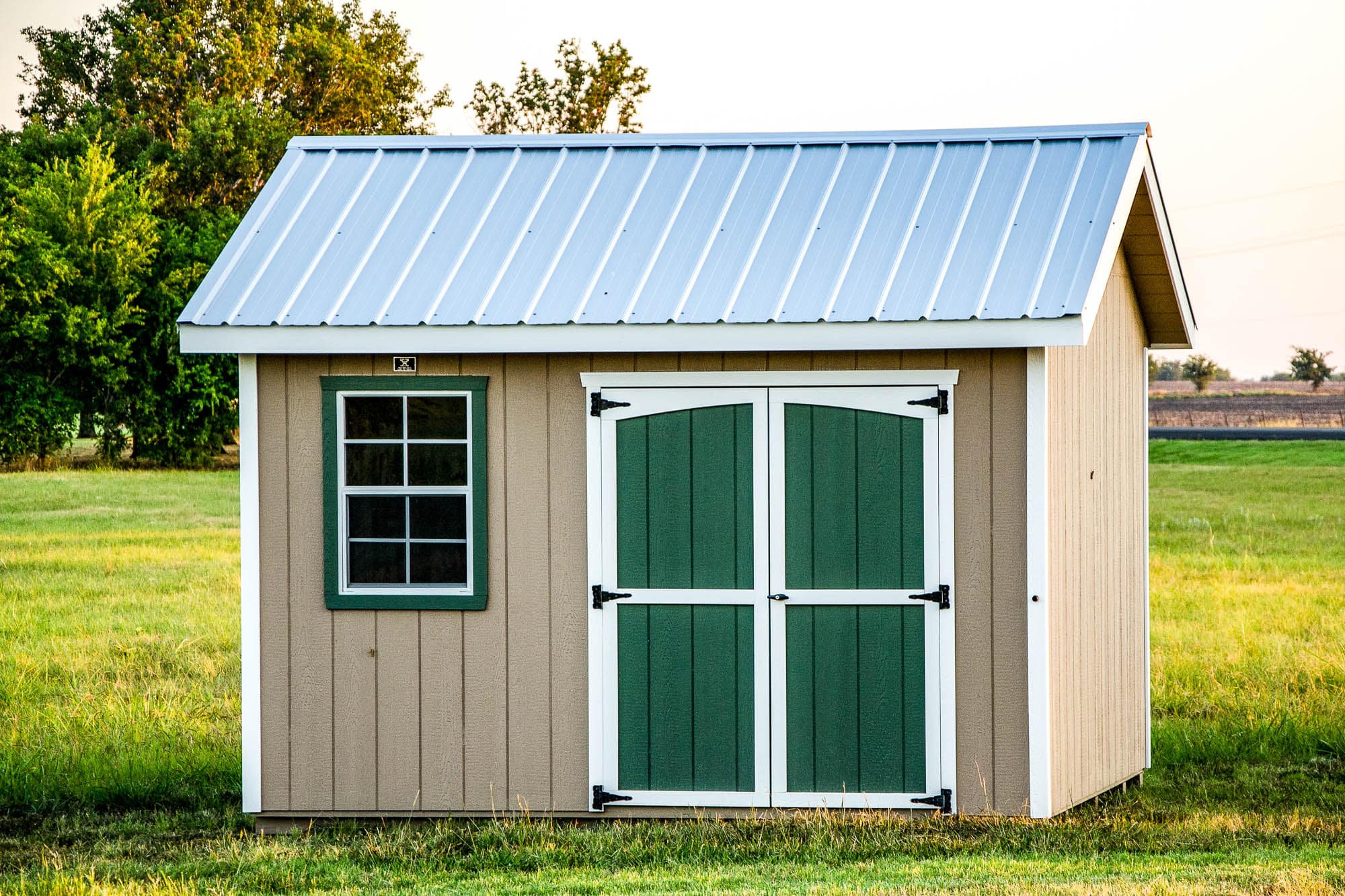 A beige classic shed with a white metal roof, green double doors with white trim, and two windows with white trim symmetrically positioned on either side of the doors. The shed is placed on a grassy lawn with trees in the background.
