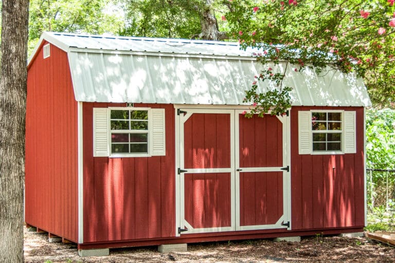A red Dutch Barn shed with a white metal roof, white trim, and two sets of double doors—one on the front and one on the side. The shed has multiple small windows with white trim, evenly placed along the sides. The structure is positioned in a shaded area with trees in the background.