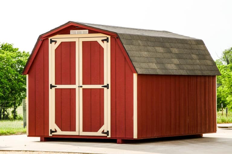 A red mini barn shed with a gambrel roof covered in dark gray shingles, beige trim, and large double doors with black decorative hinges. The shed is set on a paved surface with trees in the background.