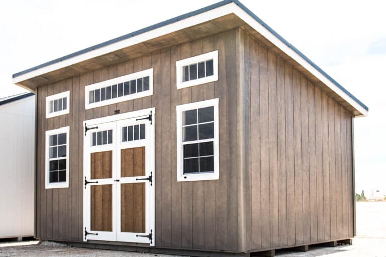 A tall, studio shed with a brown wood-grain exterior, a black metal roof, and large transom windows across the top front side. It has a set of double doors with windows, framed in white trim. The shed is positioned on a clean, level surface with trees in the background.