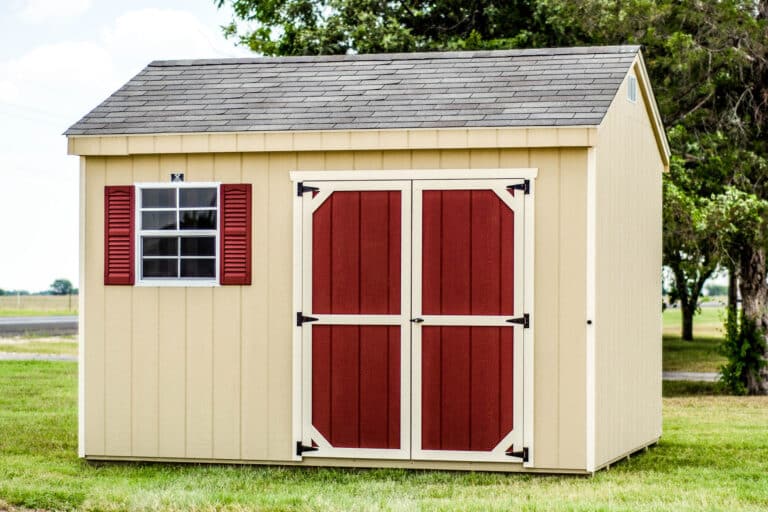 A beige workshop shed with a dark gray shingled roof, red barn-style double doors with white trim, and a small lofted window with white trim above the doors. A side window with white trim is visible. The shed is placed on a grassy lawn with trees in the background.